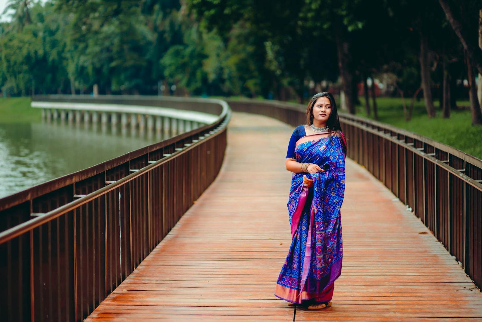 A woman in traditional sari strolls along a scenic wooden path in a lush park in Dhaka, Bangladesh.