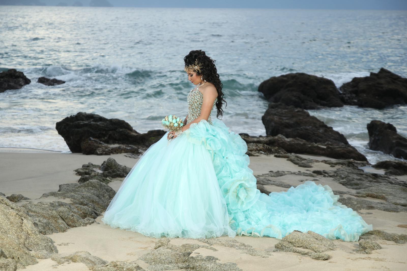 A woman in an elegant blue gown stands on a rocky beach, gazing thoughtfully at the ocean.
