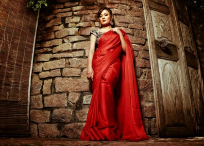 A woman in a red sari poses confidently against a stone wall with wooden doors.