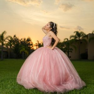 A beautiful woman in a glamorous pink gown poses elegantly in a garden at sunset.